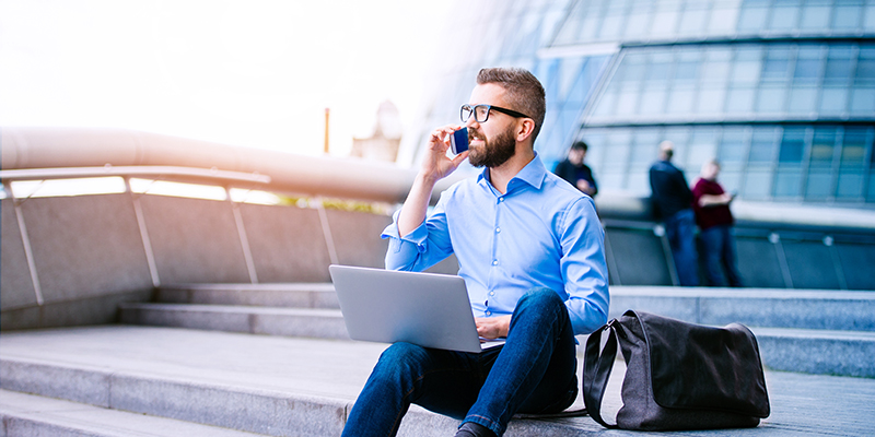 Handsome hipster manager sitting on stairs on sunny day, working on laptop, talking on a smart phone, London, City Hall